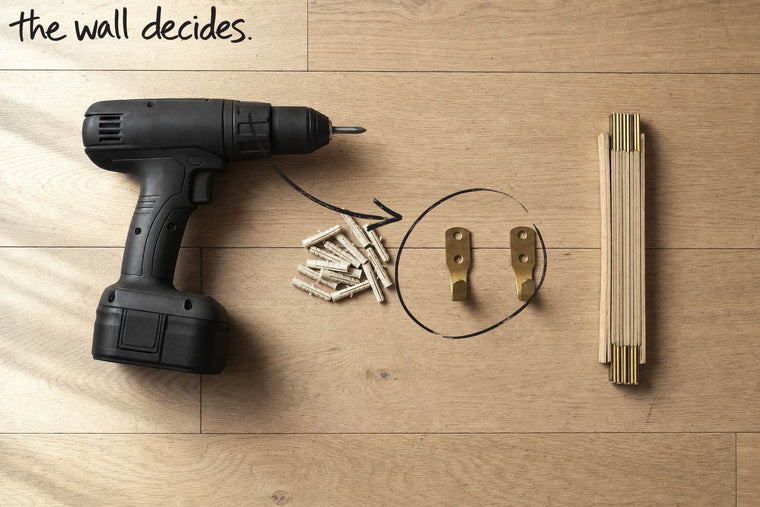 A matt black cordless drill on an oak floor with wall plugs, brass hooks and a folding rule, the brass hooks and wall plugs circled in open black marker, an arrow from the handwritten words the wall decides, by Willem van der Krieken