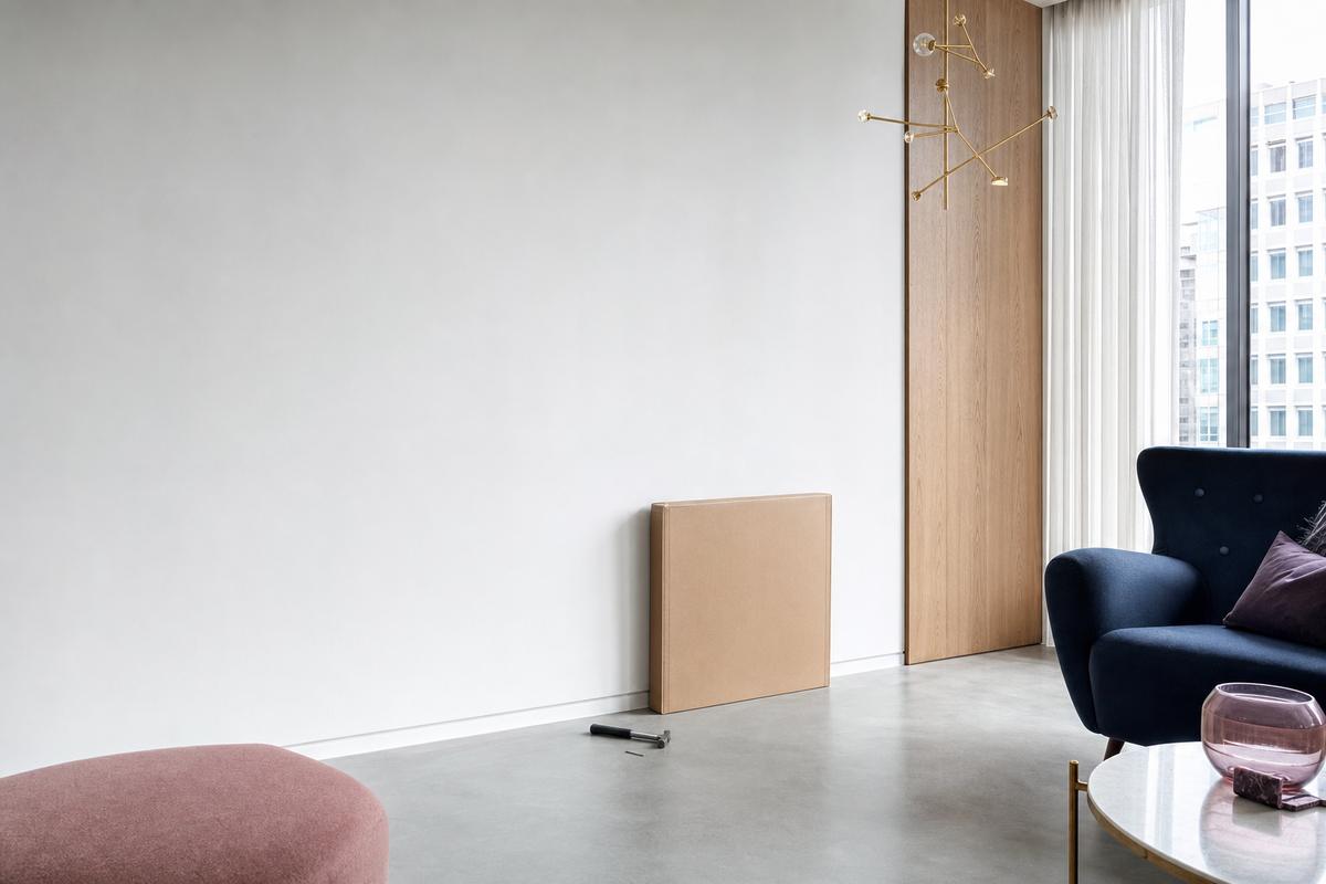 A plain unmarked cardboard box leaning against a bare pale wall in a bright hall, with a hammer and one nail on the floor beside it